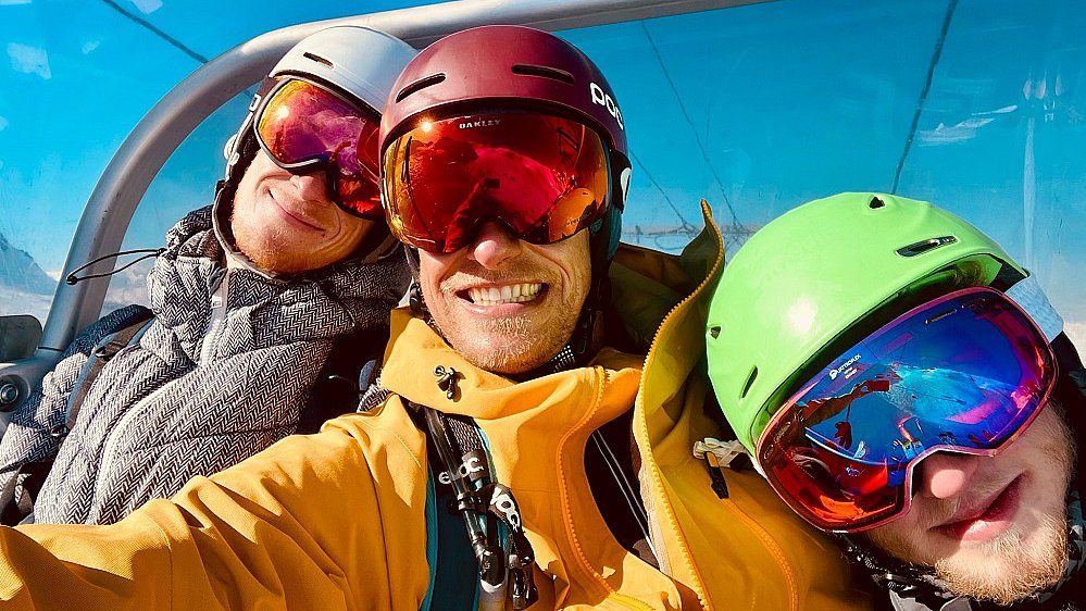 Drei Personen sitzen in einem Sessellift, sie tragen bunte Skibekleidung, Helm und Skibrille und lachen in die Kamera.