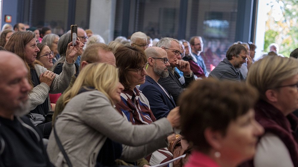 Blick in das Publikum in der Sporthalle. Zahlreiche Personen sitzen auf Stühlen und schauen nach vorne. Eine Frau filmt mit ihrem Handy.