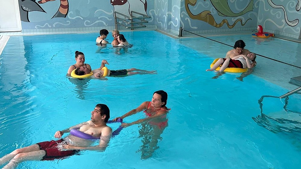 Acht Personen sind in einem Schwimmbecken mit blauem Wasser. Im Hintergrund sieht man die Wände des Schwimmbads, bemalt mit bunten Tieren. Die Personen sind jeweils zu zweit beisammen im Wasser, wobei eine Person steht und die andere Person festhält, die in einem Schwimmreifen oder in einer Schwimmnudel mit dem Rücken auf dem Wasser liegt.