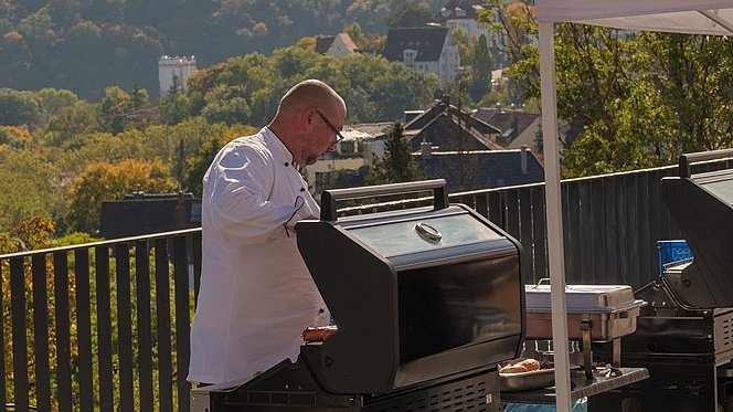 Auf der Terasse steht ein Mann in weißem Hemd hinter einem großen Grill. Hinter ihm blickt man auf Wald und Häuser.