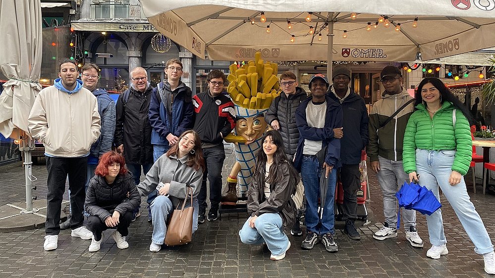 Eine gruppe junger Erwachsener steht auf der Straße unter einem weißen Sonnenschirm. Daneben steht eine Figur einer Tüte Pommes