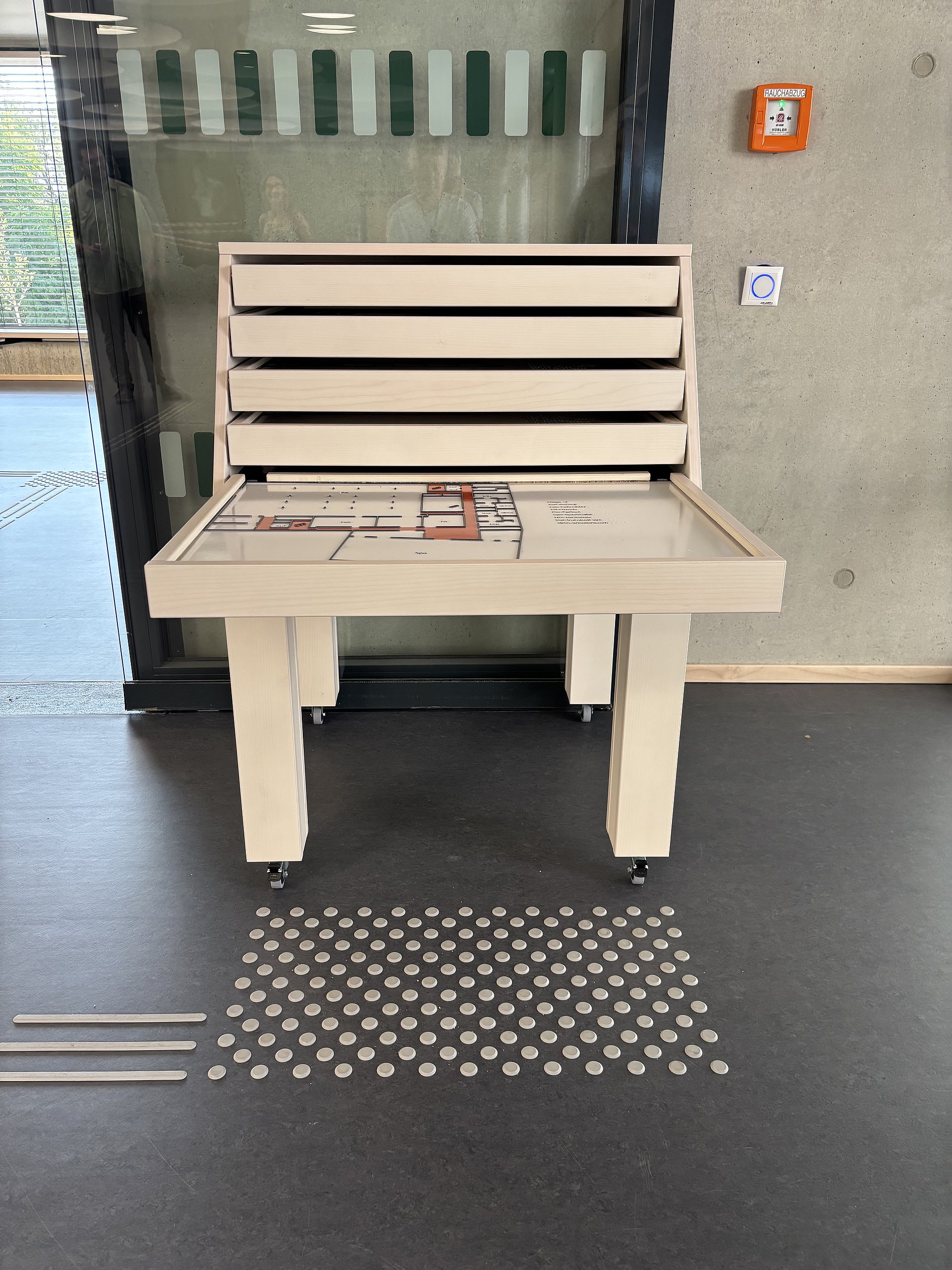 Ein hölzerner Informationstisch mit erhöhten Elementen steht auf einem dunklen Boden. Auf dem Tisch liegt eine Art Plan oder Karte mit einem markierten Bereich, vermutlich ein Gebäudegrundriss; darauf sind verschiedene Rechtecke und Wege in Weiß, Grau und Orange zu erkennen. Über der Tischfläche befindet sich ein senkrechter Aufbau mit mehreren waagerechten Leisten; sie wirken wie flache Schubladen oder Tafeln, die leicht nach vorne geneigt sind.