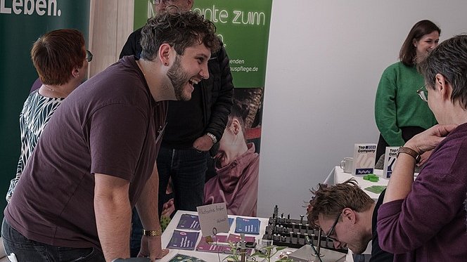 Ein junger Mann steht hinter einem großen Tisch, auf dem Flyer und Materialien ausgebreitet sind. Hinter ihm stehen zwei große Rollups der Nikolauspflege. Er lächelt und unterhält sich mit Personen, auf der anderen Seite des Tisches.