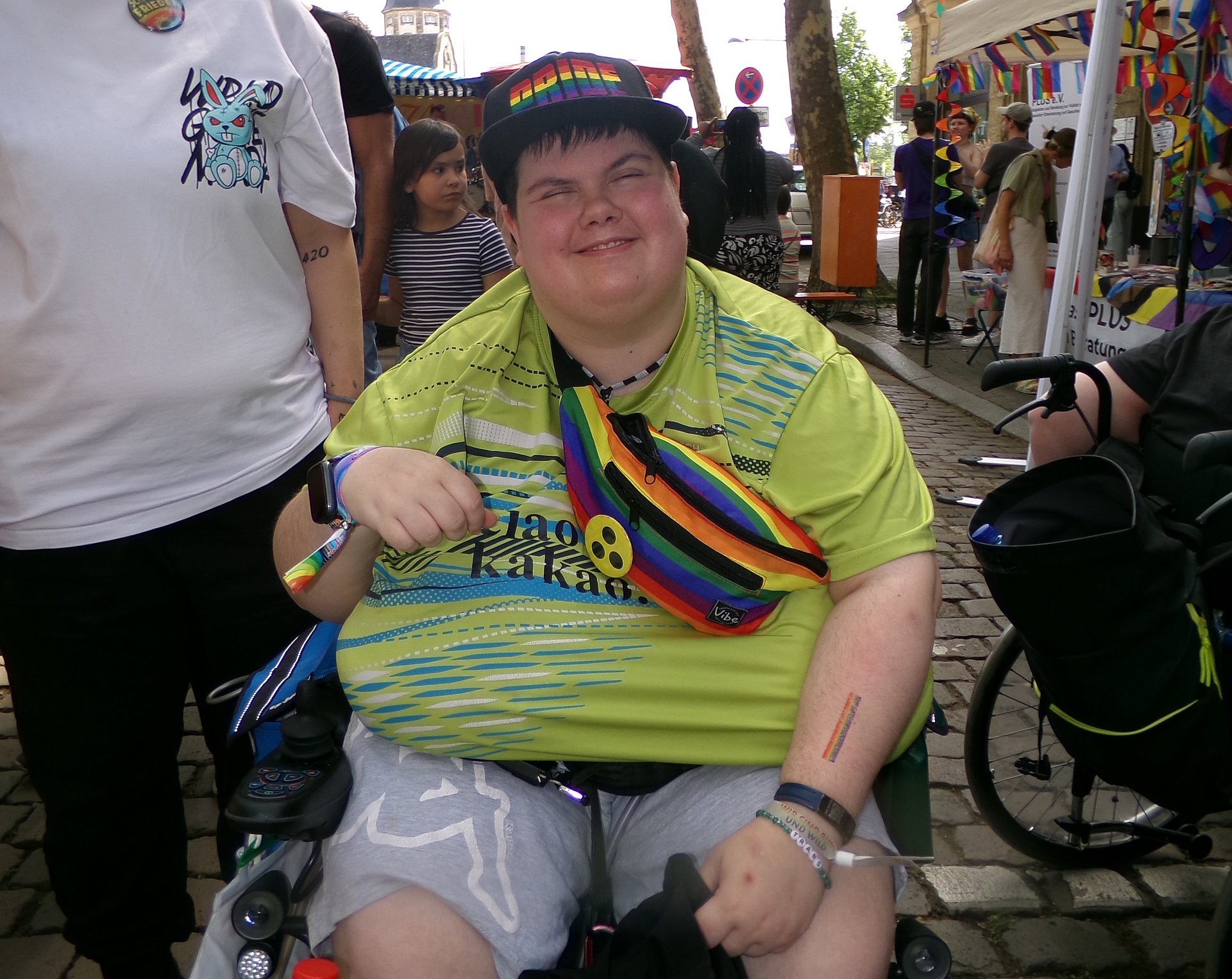 Ein Mann sitzt auf einem Rollstuhl. Er trägt ein Kappie ein kurzes T-Shirt und eine bunte Umhängetasche an der ein Blindenabzeichen zu erkennen ist. Er lacht fröhlich.
