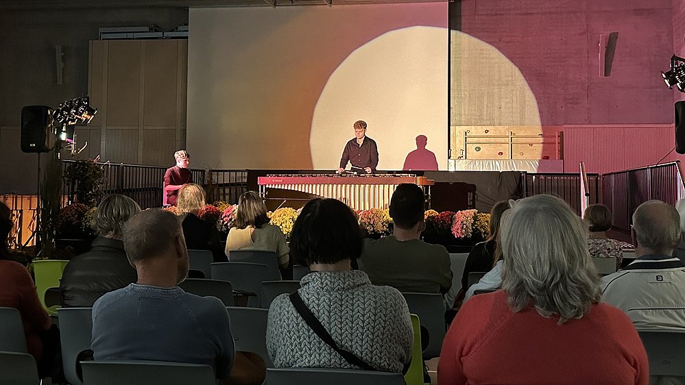 In der Sporthalle sitzen mehrere Gäste auf Stühlen. Vor ihnen auf der Bühne steht ein e große Marimba, ein junger Mann steht dahinter und spielt auf ihr. Er wird mit einem großen Lichtkegel beleuchtet.
