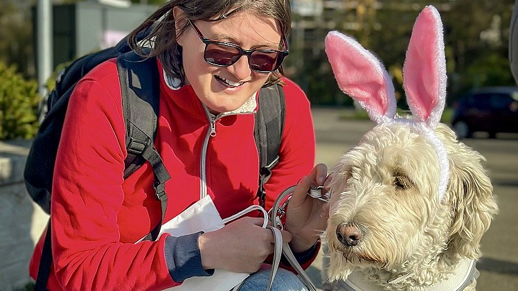 Eine Frau mit dunklen langen Haaren, roter Jacke und blauer Jeans kniet neben einem Hund mit hellem Fell. Der Hund trägt einen Haarreif mit rosafarbenen Hasenohren.