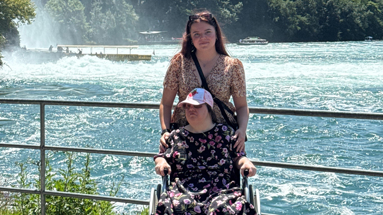 Eine Frau mit geblümtem Kleid und weißer Capie sitzt in einem Rollstuhl. Hinter ihr steht eine Frau in Bluse und mit langen dunklen Haaren, die den Rollstuhl von hinten hält. Beide Frauen schauen in Richtung der Kamera. Im Hintergrund ist ein Geländer und dahinter ein breiter Fluss mit blauem Wasser, grüne Bäume und die Gischt eines Wasserfalls.