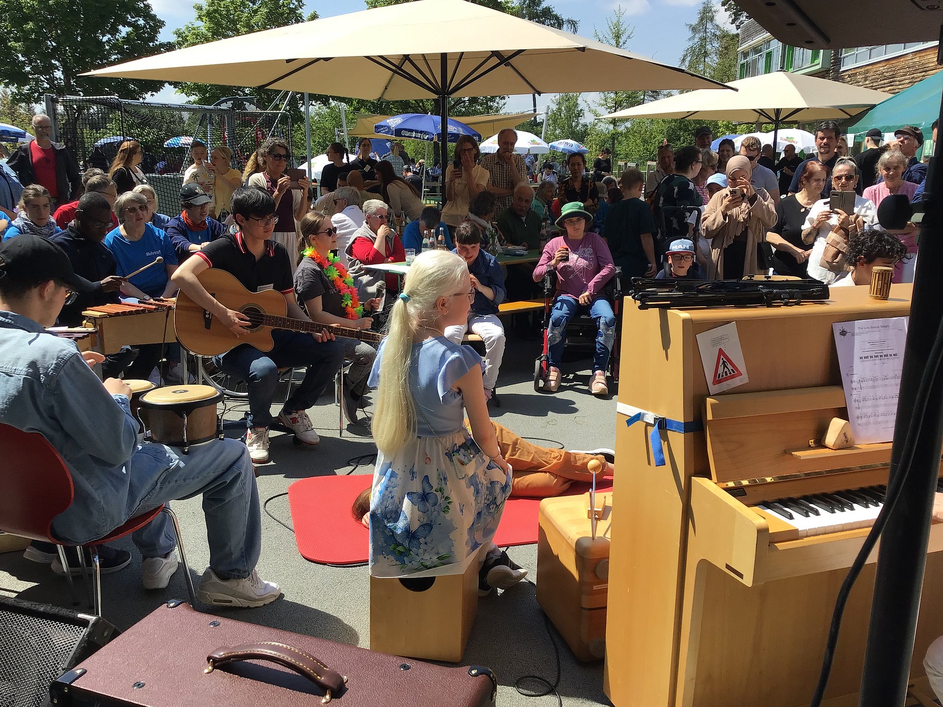 Auf dem Foto sitzt links eine Person mit blonden Haaren, rot‑weiß gestreiftem Oberteil und weißer Hose an einem hellen Holzklavier oder -orgel und spielt. Vor ihr liegt ein aufgeklapptes Notenblatt auf dem Instrument. Rechts und im Hintergrund steht und sitzt eine größere Gruppe von Menschen in sommerlicher Kleidung, die der Musik zuhören; einige tragen bunte Accessoires wie Hüte oder Blumenketten. Im unteren Bildbereich ist eine Person mit Strohhut und gemustertem Hemd von hinten zu erkennen, die ein Saiteninstrument mit rundem Korpus umgehängt hat. Im Hintergrund des Fotos sind ein Basketballkorb, Tische und Bänke sowie bunte Wimpel zu erkennen, was auf ein fröhliches Freiluftfest bei sonnigem Wetter schließen lässt.