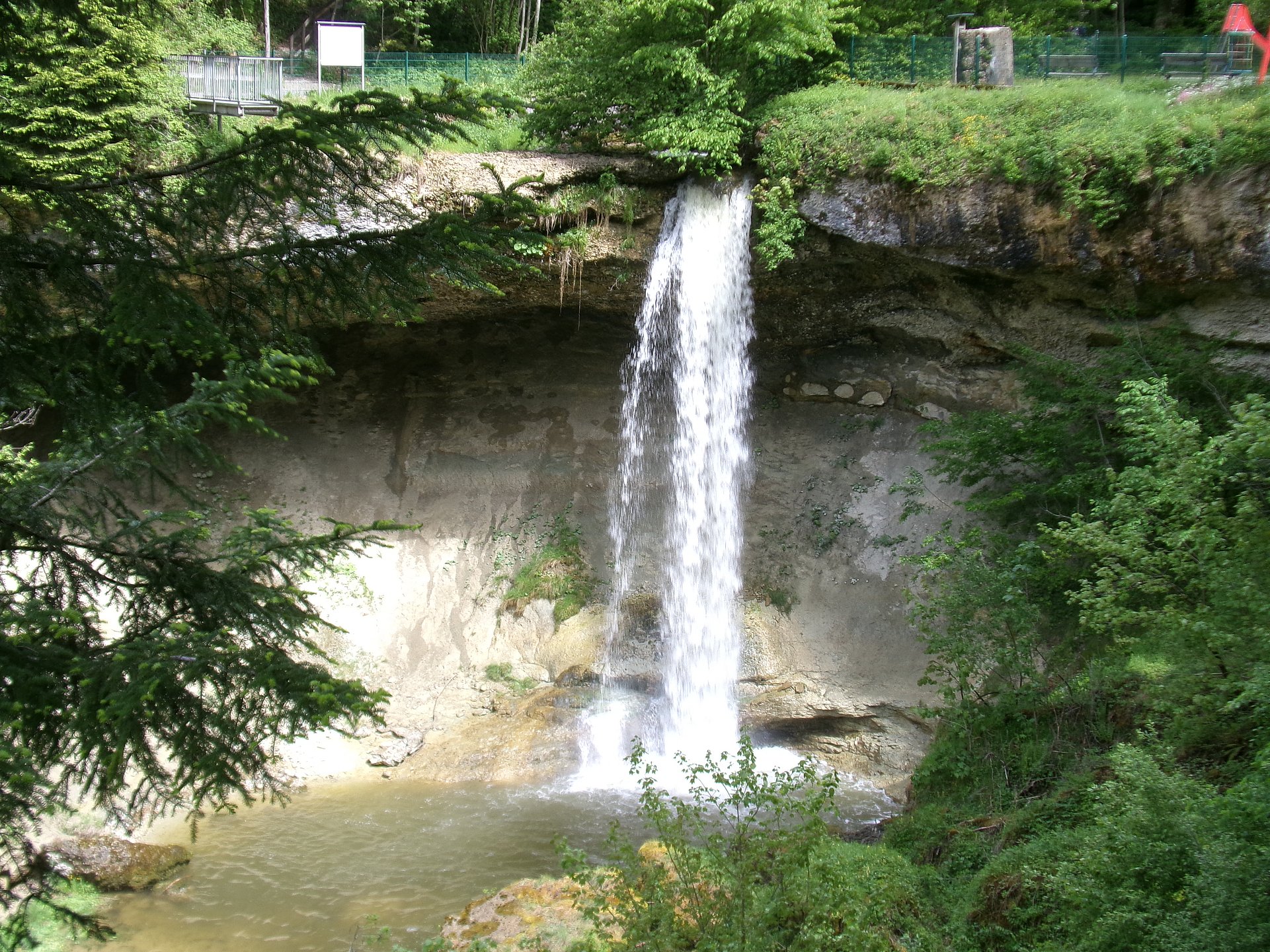 Ein Wasserfall, der von einer Felskante in ein darunterliegendes Becken stürzt.   Das Wasser fällt in einem breiten, weißen Schleier die graue Felswand hinunter