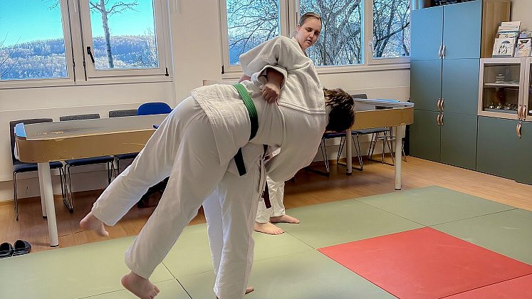 Eine junge Frau im weißen Kampfsport-Anzug hält mit der rechten Hand den Rücken einen anderen jungen Mannes im weißen Anzug, dieser fliegt quer vor ihr in der Luft.