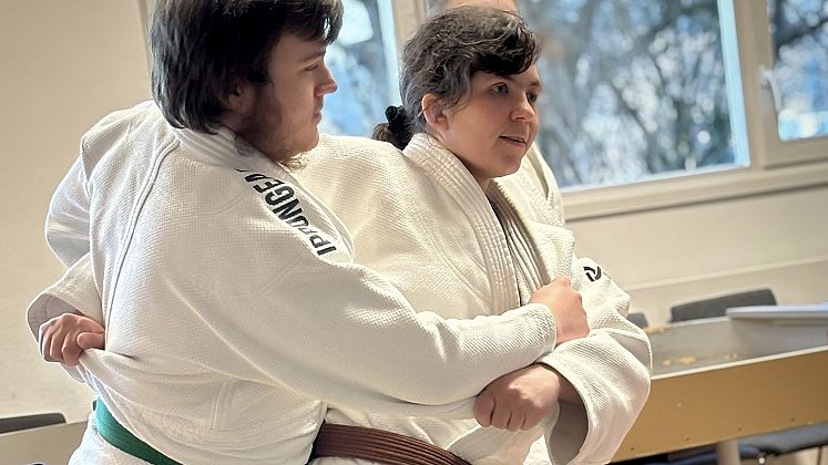 Ein junger Mann und eine junge Frau stehen schräg hintereinander. Sie tragen einen weißen Kampfsport-Anzug mit grünem bzw. braunem Gürtel. Ihre Hänge umgreifen den Bauch bzw. Rücken des jeweils anderen.