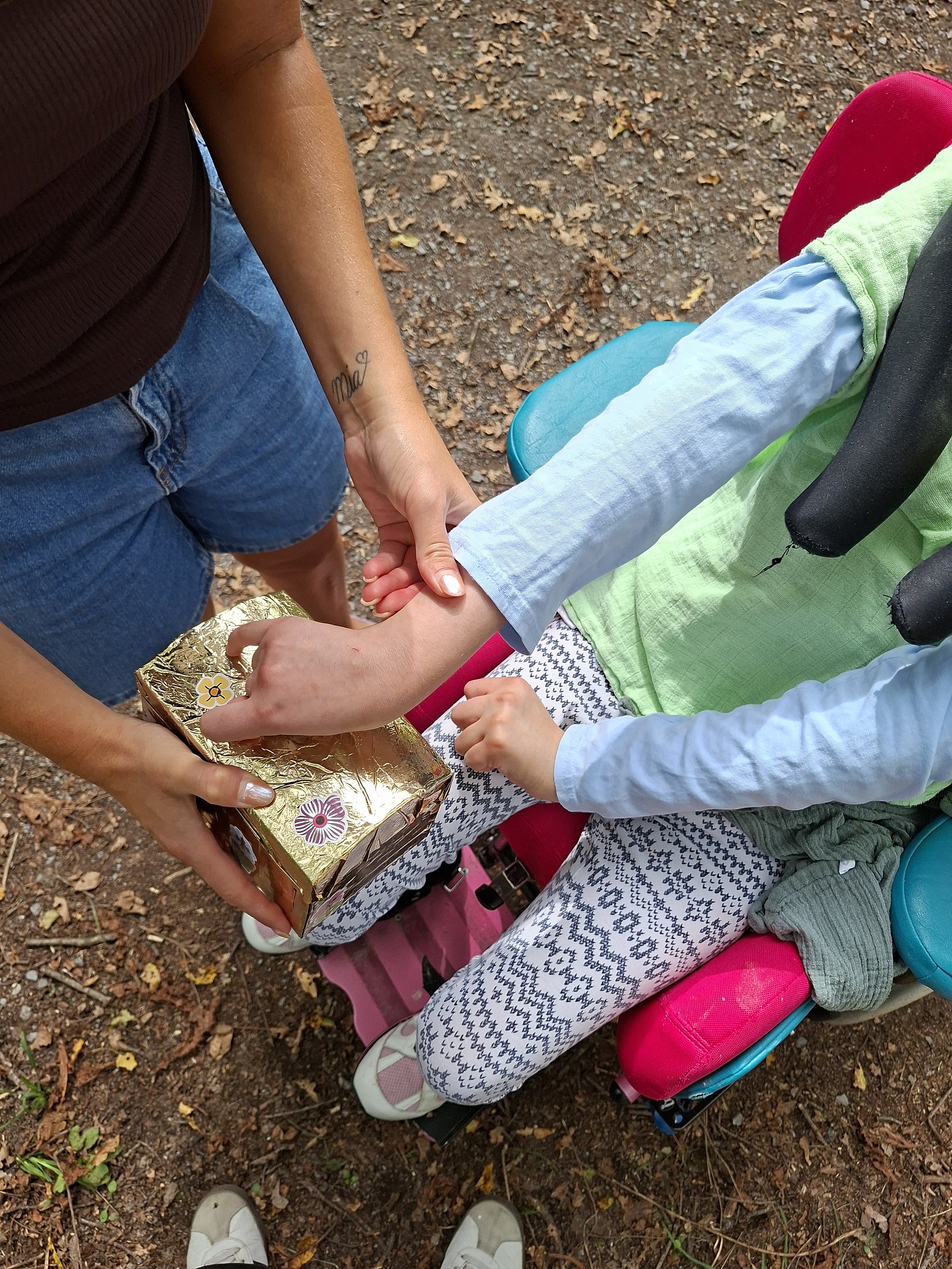 Das Foto zeigt zwei Personen im Freien auf einem Waldboden mit Laub und kleinen Ästen. Rechts sitzt ein Kind in einem bunten Rollstuhl mit pinkfarbenem Sitz mit blauen Lehnen. Das Kind trägt eine helle, gemusterte Hose, helle Schuhe und ein hellblaues Langarmshirt. Links auf dem Bild steht eine erwachsene Person mit dunkelbraunem, ärmellosem Oberteil und blauer kurzer Jeanshose. Diese Person hält mit einer Hand eine golden glänzende Schachtel mit Blumenstickern obenauf. Mit der anderen Hand führt sie die Hand des Kindes im Rollstuhl an die Schachtel heran.