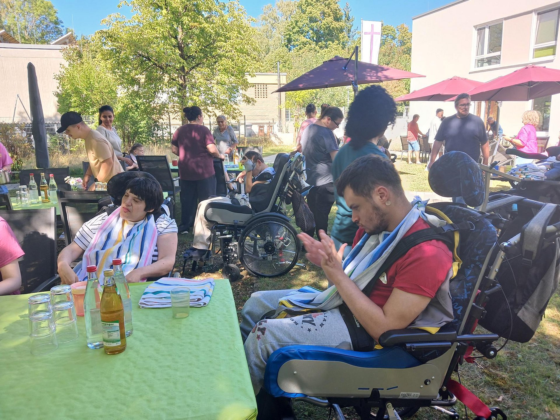 Grillfest im Freien mit vielen Menschen an Tischen und in Rollstühlen.