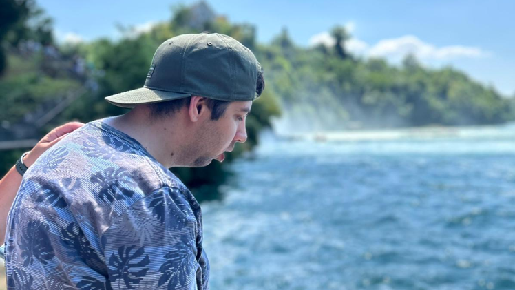 Ein Mann mit kurzen dunklen Haaren und grauen Capie hält sich mit beiden Händen an einem Geländer fest. Er trägt ein T-Shirt mit Pflanzenblatt-Aufdrucken. Vor ihm hinter dem Geländer ist ein breiter Fluss mit blauem Wasser, grüne Bäume und die Gischt eines Wasserfalls.