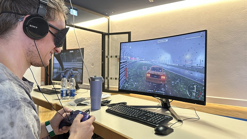 Ein junger Mann mit Headset an einem Gaming Platz. Er hat kurze, dunkelbraune Haare, hellen Hauttyp und trägt ein graues T Shirt. Über den Ohren sitzen schwarze, ohrumschließende Kopfhörer von Sony, vor dem Mund ein Mikrofonbügel. Auf den Augen trägt er eine schwarze Maske. Er hält mit beiden Händen einen Gamecontroller, der mit einem Kabel verbunden ist. Am Handgelenk ist ein grünes Stoffbändchen. Vor ihm steht auf dem Tisch ein Monitor, auf dem ein Rennspiel läuft: Ein rotes Auto fährt bei Regen auf einer Straße, es sind Regentropfen auf der „Kamera“ und eine nasse Fahrbahn zu erkennen. Unten links auf dem Bildschirm ist eine runde Anzeige, vermutlich Tacho oder Minikarte, oben rechts ein kleines Textfeld.