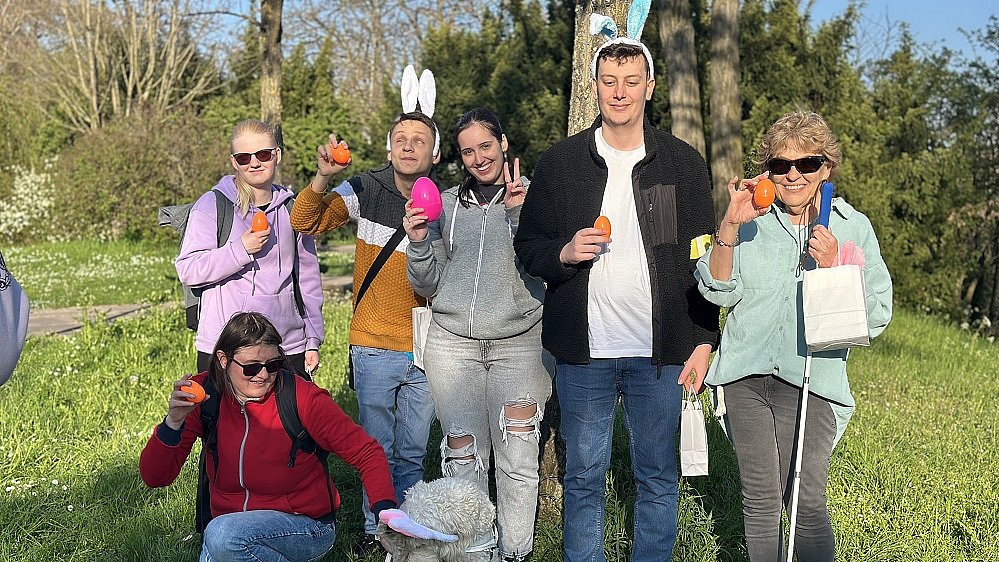 Sechs junge Menschen und Erwachsene stehen nebeneinander und schauen in die Kamera, zwei von ihnen tragen einen Haarreif mit großen Hasenohren, viele halten bunte Eier in die Höhe. Vor ihnen sitzt ein Hund mit hellem Fell.