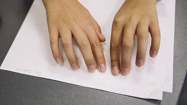 Zwei Hände tasten mit den Fingern Braille-Schrift auf einem Blatt Papier