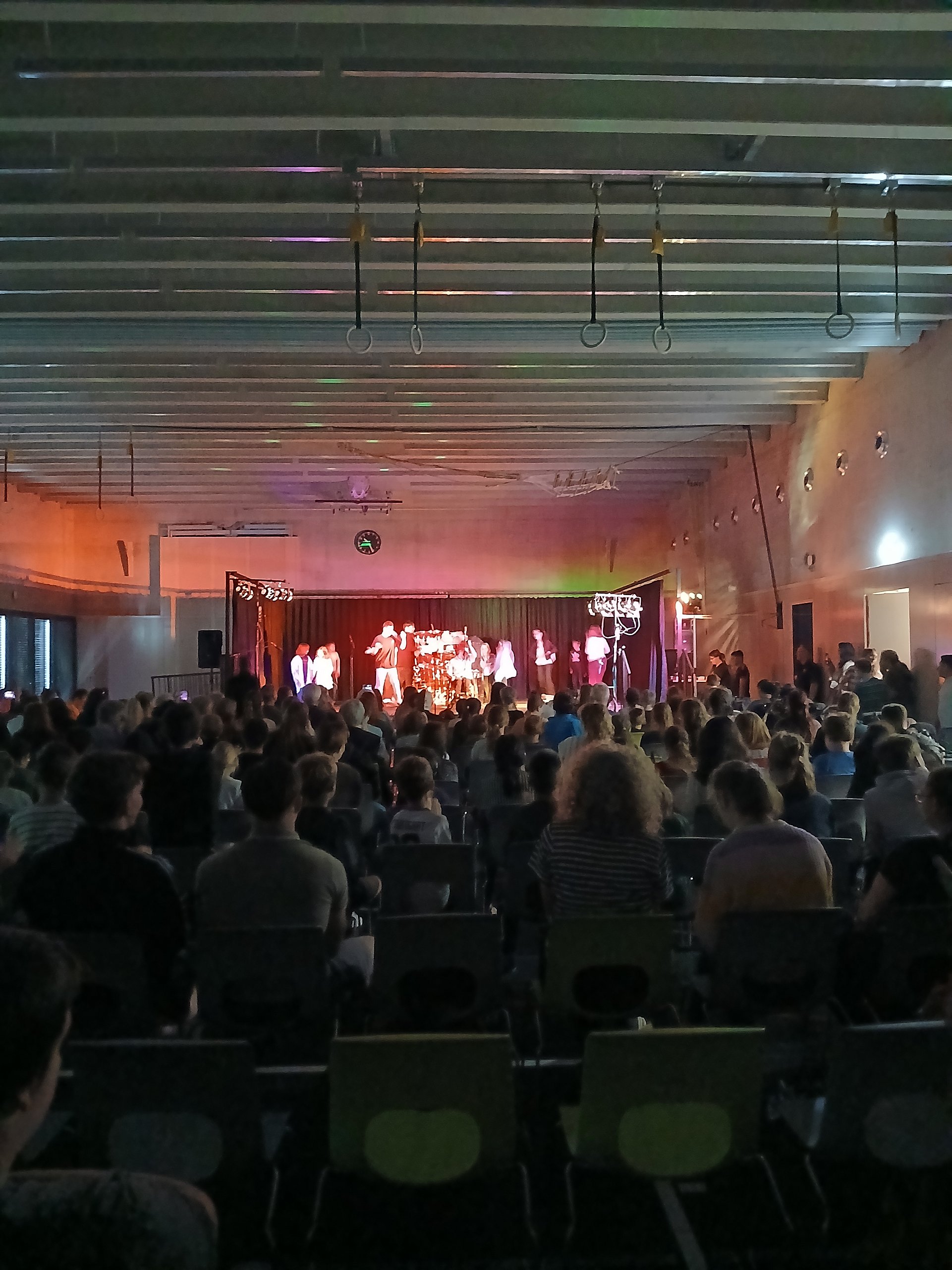Ein großer Saal mit vielen Stuhlreihen, die auf eine Bühne ausgerichtet sind. Auf den Stühlen sitzt ein dichtes Publikum, überwiegend Kinder, Jugendliche und einige Erwachsene, teils mit hellen, teils mit dunklen Haaren, viele mit Freizeitkleidung wie T‑Shirts und Kapuzenpullis. Vorne befindet sich eine erhöhte Bühne mit rotem Vorhang und farbigem Bühnenlicht in Rot‑ und Violetttönen. Auf der Bühne stehen mehrere Personen nebeneinander, vermutlich eine größere Gruppe, deren Gesichter wegen der Entfernung nicht klar erkennbar sind; sie tragen unterschiedliche, eher helle und bunte Kleidung. Links und rechts der Bühne sind Lautsprecher und Lichtständer aufgebaut, rechts steht ein beleuchteter Kronleuchter‑Dekorationsständer. Über der Bühne hängt eine analoge Uhr an der Wand. Die Decke des Saals ist mit länglichen Metallstreben und mehreren Ringen oder Halterungen versehen, von denen einige herabhängen. Die Wände rechts und links sind schlicht, mit kleinen, runden Wandleuchten, die warmes Licht geben.