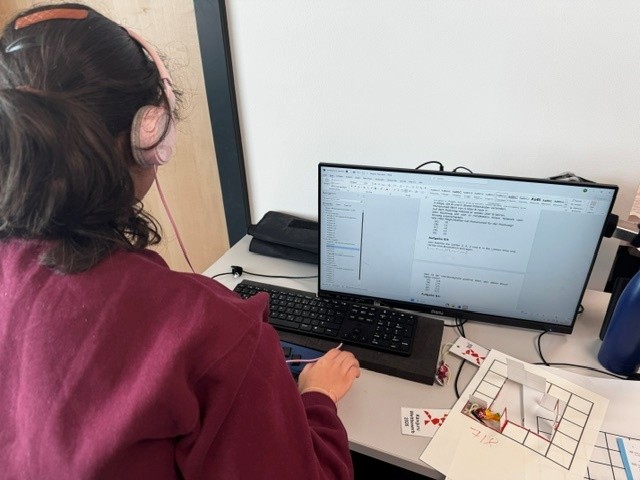 Eine Person mit dunkelbraunen, zusammengebundenen Haaren und heller Haut sitzt an einem Schreibtisch und arbeitet an einem Computer. Die Person trägt einen bordeauxroten Pullover und ist seitlich von hinten zu erkennen, über die Tastatur gebeugt. Vor ihr steht ein breiter, hochkant gedrehter Monitor, auf dem eine Programmier- oder Entwicklungsumgebung mit mehreren Fenstern geöffnet ist; in den Fenstern sind Tabellen und längere Textblöcke zu sehen, aber die Schrift ist zu klein, um sie lesen zu können. Unter dem Monitor liegt eine schwarze Tastatur, rechts daneben ein schwarz-graues Laptop, das halb unter dem Monitor steht.