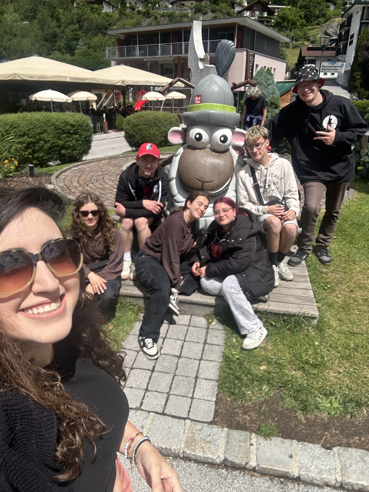 Selfie-Gruppe im Freien mit einer großen Comic-Figur in der Mitte. Im Vordergrund links ist ein lachendes Gesicht einer Frau mit langen lockigen braunen Haaren, großer Sonnenbrille und schwarzem Oberteil, der Unterarm mit Armbändern ist zu sehen, sie hält die Kamera schräg von oben. Hinter ihr sitzt und kniet eine Gruppe von sechs jungen Menschen auf einem gepflasterten Weg und auf einer niedrigen Holzbank, nah beieinander um eine große graue Tier-Figur herum.