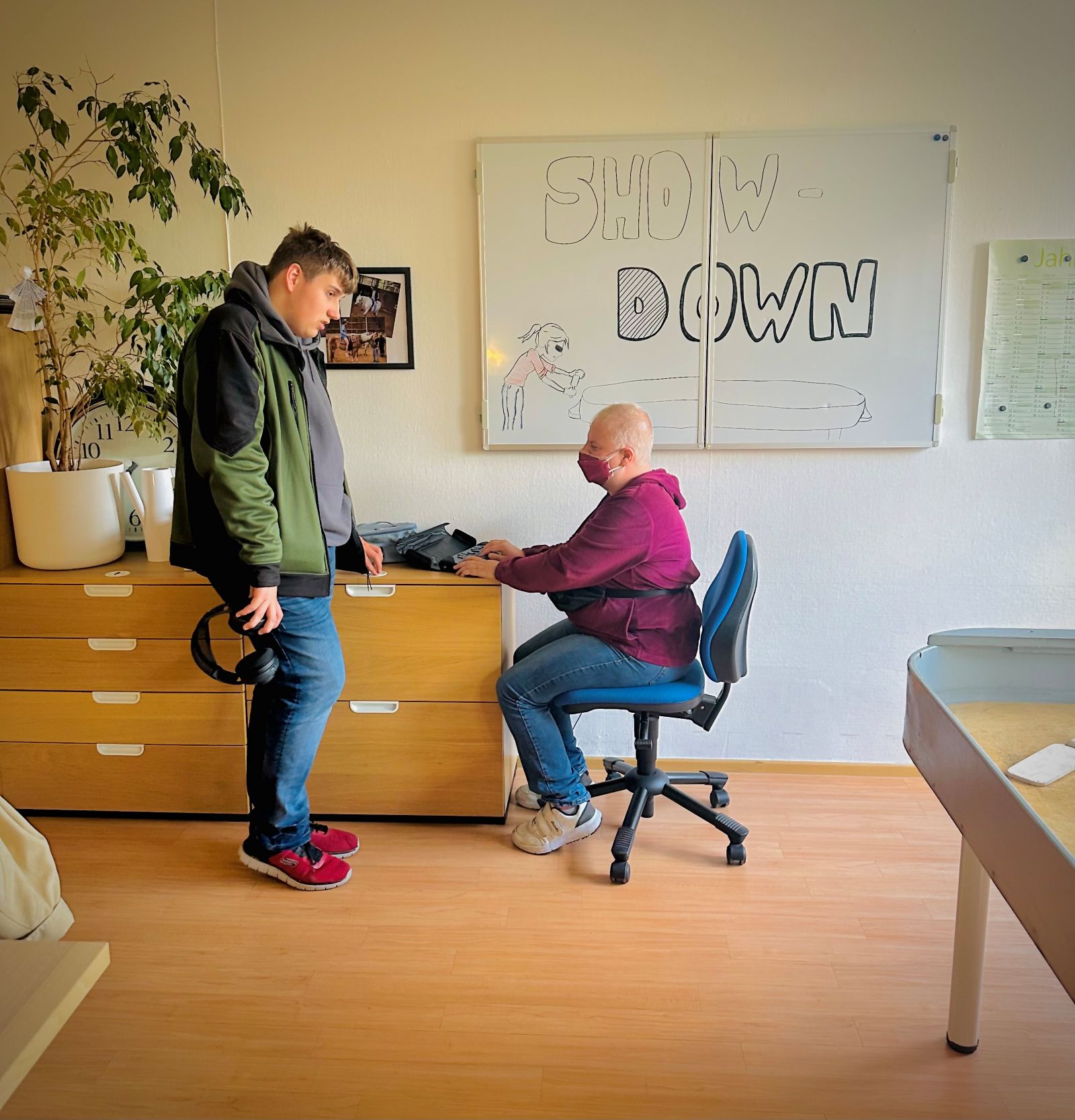 Ein junger Mann steht neben einer Kommode. Dort sitzt ein Mann auf einem Drehstuhl und tippt etwas in seine Punktschriftmaschine. Hinter ihm steht auf einem großen Plakat an der Wand Show Down