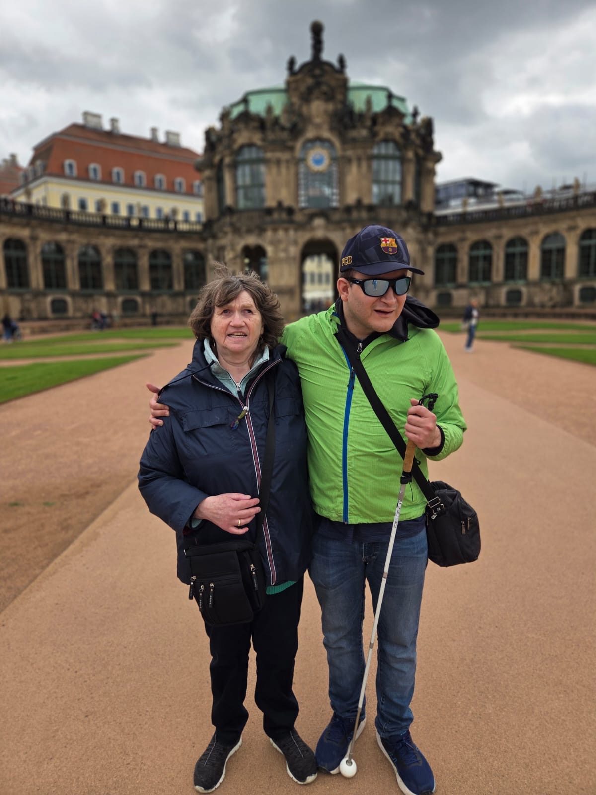 Zwei Personen stehen eng umarmt auf einem breiten sandfarbenen Weg vor einem historischen Gebäude. Links steht eine Frau mittleren Alters mit heller Haut und mittelbraunen, leicht gewellten, kinnlangen Haaren. Sie trägt eine dunkelblaue Outdoor-Jacke mit hellblauem Futter, darunter ein helles Oberteil, eine dunkle Hose und schwarze Sportschuhe mit weißer Sohle. Über der Schulter hängt eine schwarze Umhängetasche, die seitlich vor ihrem Oberschenkel liegt. Ihr linker Arm hängt locker, mit der rechten Hand hält sie sich am Rücken der anderen Person fest und lächelt.